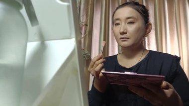 Asyalı bir kadının aynanın karşısına fırçayla göz farı sürerken yakın plan fotoğrafı. Taze, ışıl ışıl bir görünüş için günlük güzellik..
