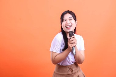 Neşeli bir Asyalı kadın mikrofonu tutarken gülümsüyor, başka tarafa bakıyor, gösterisine hazır. turuncu arkaplanda izole