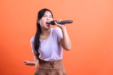 Hevesli genç bir kadın, turuncu arka planda izole edilmiş saf neşe ve enerjiyi ifade eden bir mikrofonla şarkı söylüyor.