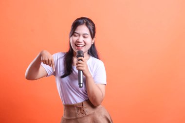 Genç bir kadın mikrofona şarkı söylerken geniş bir gülümseme sergiliyor ve parmağını turuncu arka planda soyutlanmış bir şekilde aşağıya doğru işaret ediyor.