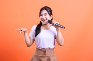 Mikrofonla şarkı söyleyen ve stüdyoda sol eliyle işaret eden genç bir kadın çok mutlu ve arkadaş canlısı görünüyor. turuncu arkaplanda izole