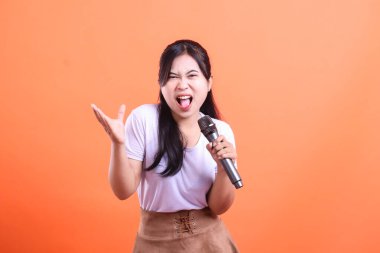 Mikrofonlu genç ve etkileyici bir kadın, performansı sırasında turuncu arka planda izole edilmiş canlı bir duygu ile tutkuyla şarkı söylüyor.