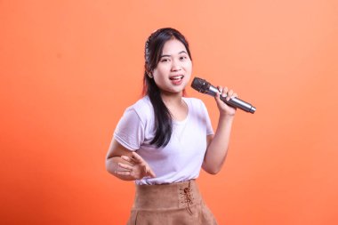 Mikrofonlu genç bir kadın, tutkuyla şarkı söylüyor, gülümsüyor ve performansından zevk alıyor. turuncu arkaplanda izole