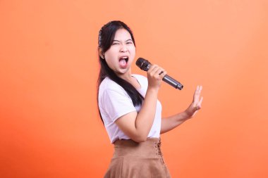Mikrofonlu genç bir kadın tutkuyla şarkı söylüyor. Ağzı açık, eli havada ve anlamlı bir yüzü var. Turuncu arka planda soyutlanmış..