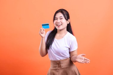 Neşeli genç bir kadın, neşeli bir ifade ve çekici bir poz sergileyen kredi kartını hevesle sunar. turuncu arkaplanda izole