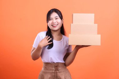 Neşeli Asyalı bir kadın karton kutuları taşırken gülümsüyor. Sevkiyat ya da teslimata hazır. turuncu arkaplanda izole