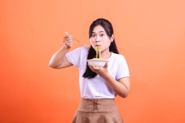 Şaşırmış bir ifadeyle genç bir kadın beyaz bir kaseden çatalla erişte yiyor. turuncu arkaplanda izole