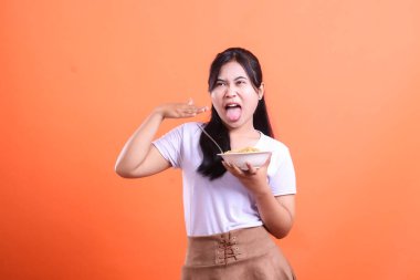 Genç bir kadın elinde bir kase erişte tutarken tiksinti ifadesi gösteriyor. Mutsuz ve üzgün görünüyor çünkü yemeklerden hoşlanmıyor, turuncu arka planda izole edilmiş.