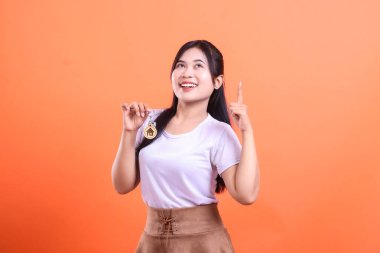 Elinde anahtar tutan gülümseyen bir kadın, işaret parmağıyla yukarı bakıyor, sanki bir fikri varmış ya da heyecan gösteriyormuş gibi; turuncu arka planda izole edilmiş.