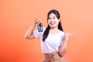 Araba anahtarı tutan, gülümseyen ve bir şey göstermek için baş parmağını gösteren genç bir kadın. turuncu arkaplanda izole