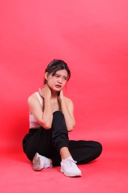 Asya 'lı bir kadın kırmızı arka planda siyah spor ayakkabı ve pantolon giyerken ağrıyan boynunu tutarak oturur..