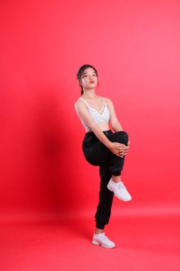 Siyah pantolonlu ve beyaz bluzlu bir kadın, elleri dizine yakın, kırmızı bir arka planda izole bir şekilde bacağını uzatır..
