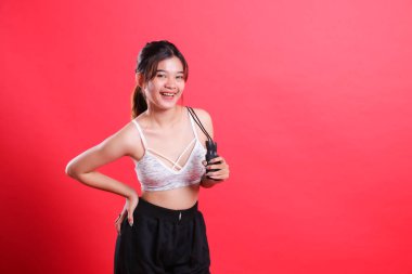 Gülümseyen bir kadın elinde atlama ipi tutuyor, spor sutyen ve siyah şort giyiyor, kırmızı arka planda izole edilmiş..