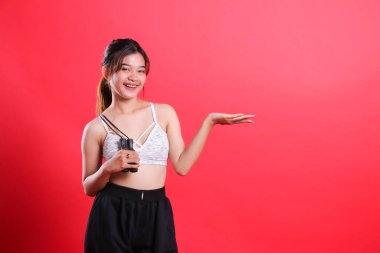 Beyaz, siyah bluzlu ve siyah pantolonlu bir kadın atlama ipini tutuyor ve diğer eliyle kırmızı arka planda izole bir şekilde boş bir alana sunuyor..