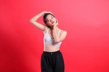 Beyaz spor bluzlu ve siyah pantolonlu bir kadın elleriyle boynunu uzatıyor. Kırmızı bir arka planda izole edilmiş..