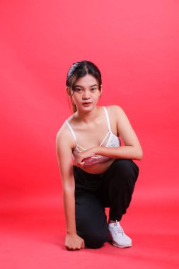 Beyaz sutyen ve siyah pantolon giyen bir kadın, kameraya bakarken çömeliyor, kırmızı arka planda izole edilmiş.