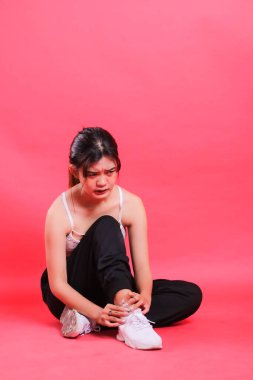 Kırmızı arka planda, siyah spor ayakkabı ve pantolon giyerken yaralı bileğini acı içinde tutan bir kadın oturuyor..