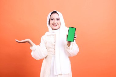 tesettürlü tesettürlü gülümseyen Müslüman kadın elinde yeşil ekranla akıllı telefon tutuyor ve telif hakkı için el hareketi yapıyor. turuncu bir arkaplanda izole.