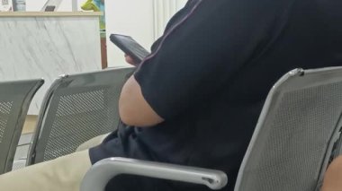 Bu görüntü, bekleme salonunda otururken akıllı telefon kullanan birini gösteriyor. Bireyin cihaza odaklanması, hizmet beklerken eğlence ve iletişim için teknolojiye güvenmeyi gerektiriyor.