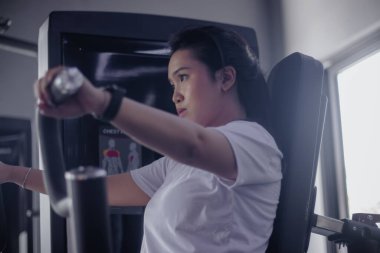 Genç bir kadın spor salonundaki bir makineyle göğüs egzersizine odaklanır. Kararlıdır ve kendini fiziksel hedeflerine adamıştır. Kapalı alanda egzersiz yapar ve saat takar..