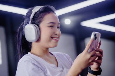 Gülümseyen genç bir kadın akıllı telefonuyla iletişim kurarken kablosuz kulaklıkla müzik dinlemekten hoşlanır. Modern bağlantıyı, rahatlığı ve teknolojiden anlayan ev yaşamını somutlaştırıyor..