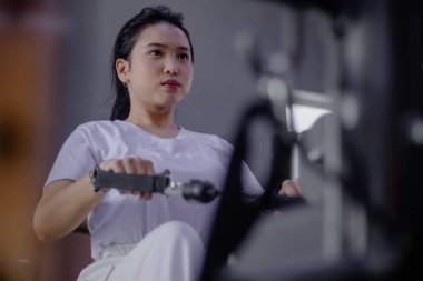 Asyalı bir kadın kendini bir fitness merkezinde kürek çekme makinesinde çalışmaya adamış. Odaklanması ve kararlılığı, sağlığına ve sağlığına olan bağlılığını gösteriyor..