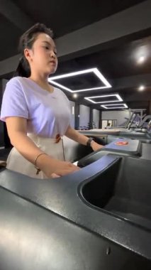 Genç Asyalı bir kadın, modern bir spor salonunda koşu bandı kullanırken antrenmanına odaklanıyor. Sağlıklı ve kararlı bir yaşam tarzının portresi..