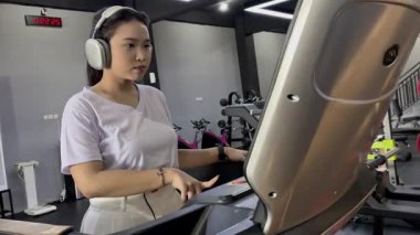 Kulaklık takan odaklanmış genç bir kadın modern kapalı bir spor salonunda koşu bandında egzersiz yapıyor. Sağlıklı bir yaşam tarzının peşinde koşmak için spor rutiniyle meşgul..