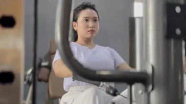 Atletik bir genç kadın modern bir spor salonunda göğüs pres makinesi kullanırken odaklanır. Fitness hedeflerine ulaşmak ve fiziksel gücünü arttırmak için çok çalışıyor..