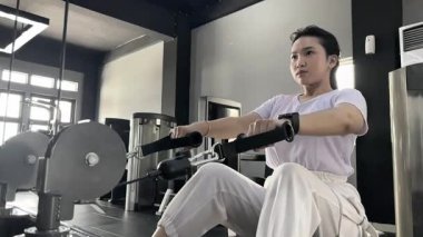 Odaklanmış Asyalı bir kadın, ışık dolu bir spor salonunda kablolu yayında egzersiz yapıyor, kararlılık gösteriyor, güç eğitimi alıyor ve sağlıklı ve aktif bir yaşam tarzına bağlı..