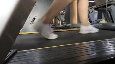 Koşu bandında egzersiz yapan bir kadının ayağını beyaz spor ayakkabılarla yakalıyor. Hareket, onun modern bir spor salonunda koştuğunu ya da hızlı yürüdüğünü gösteriyor..