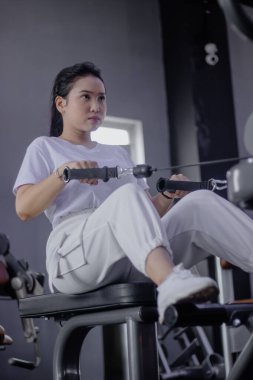 Odaklanmış genç bir kadın spor salonunda oturarak egzersiz yapıyor güçlü bir sırt egzersizi için kolu çekiyor. Sağlıklı yaşam tarzını ve fiziksel kondisyonunu teşvik etmek.