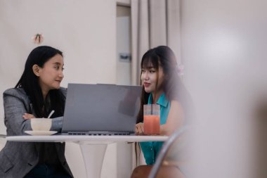 İki Asyalı kadın bir dizüstü bilgisayarda çalışıyor. Atmosfer işbirliğini ve yeniliği teşvik eder. Projelerine odaklanmış gibi görünüyorlar, lezzetli içecekler içiyorlar..