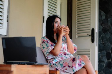 Çiçekli bornozlu kadın dışarıda laptopunun yanında sıcak bir içecek yudumluyor. Rahatlamayı uzak çalışmayla harmanlamak sakin ve üretken bir yaşam tarzı yaratıyor..