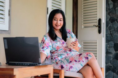 Çiçekli bir sabahlıkla kahvenin tadını çıkarırken dizüstü bilgisayarıyla uzaktan çalışan bir kadın. Mutlu kadın evde çalışıyor, özgürlüğün ve dijital göçebe yaşam tarzının tadını çıkarıyor..