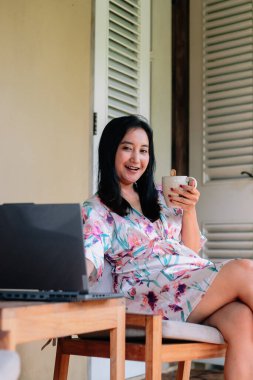 Çiçekli bornozlu, verandada kahve ve dizüstü bilgisayarın keyfini çıkaran neşeli genç bir kadın. İşi eğlence ve rahatlıkla birleştiriyor, mutlu ve neşeli görünüyor..