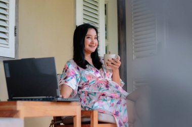 Gülümseyen bir kadın dışarıda laptopuyla çalışırken kahveden hoşlanır. Rahatlamış ve neşeli, çiçekli bir bornoz giyiyor, modern iş hayatını somutlaştırıyor..