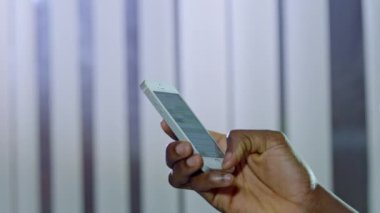 Penceredeki bulanık perdelere karşı modern cep telefonu tutan Afrikalı Amerikalı adamın eli. Kullanıcı sosyal ağda iletileri yakın planda okur