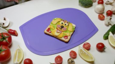Taze sebze ve peynirli gülümseyen köpek şeklinde sağlıklı bir sandviç. Salata, domates ve menekşe kesim tahtasında sosis.