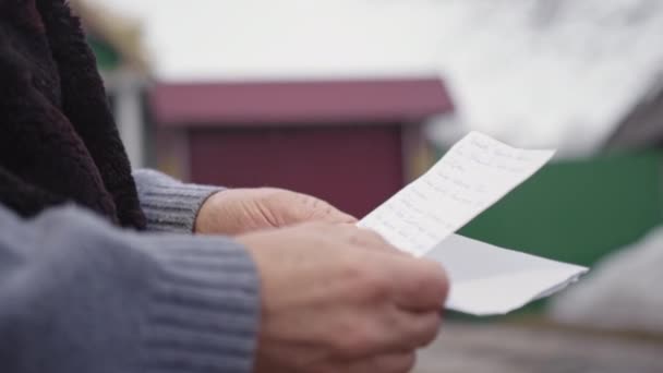 老人は田舎の庭に立つ孫娘からの手紙を読む 背景にぼやけた紙シートを持った年金受給者の手が近づく — ストック動画