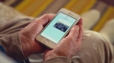 Yaşlı kadın, kocanla koltukta otururken çekilmiş fotoğraf mesajlarını kontrol ediyor. Kırışık eller arka plandaki bulanık görüntüde akıllı telefonu tutuyor.