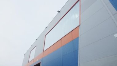 Kışın gri bulutlu gökyüzü altında endüstriyel üretim binası. Ön duvar mavi ve turuncu renklerle dekore edilmiş ve polikarbonat panelleri düşük açılı çekimde.