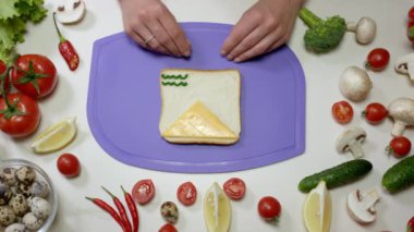 Kadın bir dilim peynir ve sebze koyar ve kesme tahtasına sağlıklı sandviç yapar. Ev hanımı menekşe doğrama tahtasında sandviç hazırlıyor.