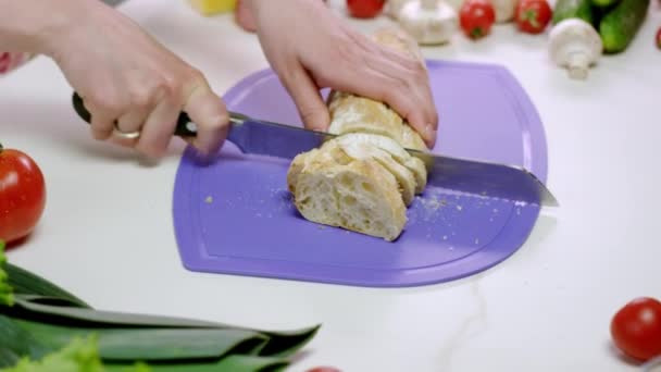 主婦はキッチンのまな板に特定のナイフで新鮮なパンをカットします 女性は野菜とバゲットでサンドイッチを作りたい — ストック動画