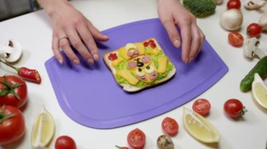 Ev hanımı kesme tahtasında gülümseyen köpek şeklinde sebzeli sandviç gösteriyor. Kadın peynir ve salatalı sandviç yapıyor.