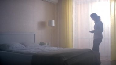 ALMATY, KAZAKHSTAN - 14 Eylül 2020: bayan odanın diğer tarafına gider ve rahat bir yatakta oturur. Kadın akıllı telefona heyecanlı bir şekilde bakıyor ve aletini yatağa koyuyor. Kadın yorgun düşmüş yatağa.