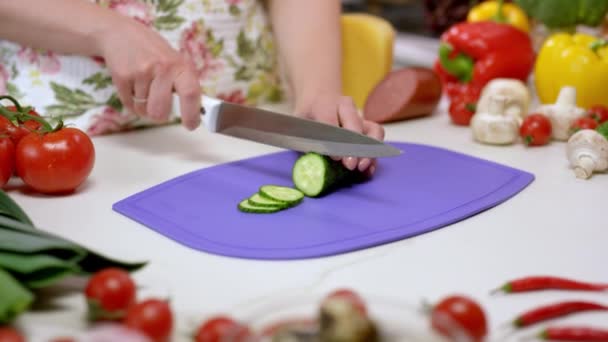 主婦はキュウリを紫色のまな板の上の薄いスライスに切ります 女性は健康的なサラダのために野菜を切るために鋭いナイフを使用します — ストック動画
