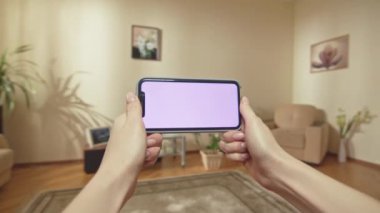 ALMATY, KAZAKHSTAN - 14 Eylül 2020: kahverengi saçlı kadın hafta sonunu akıllı telefonuyla kendi dairesinde geçiriyor. Bayan gülümsüyor ve evde oturup online video izlemekten hoşlanıyor.