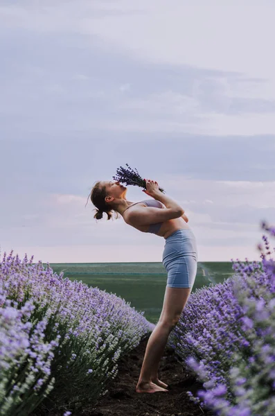 Woman in active wear doing backbend smelling lavender bouquet in lavender field