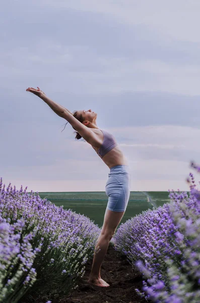 Woman in activewear doing yoga pose backbend in the middle of a lavender field on a cloudy day.